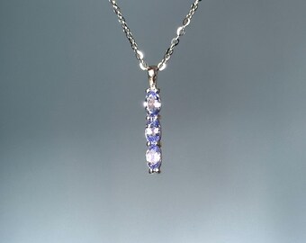 Puede incluir: Un collar de plata con un colgante vertical con tres piedras preciosas ovaladas de color púrpura claro. La cadena es delicada y el colgante está centrado, creando un diseño simple pero elegante. Las piedras están espaciadas uniformemente.