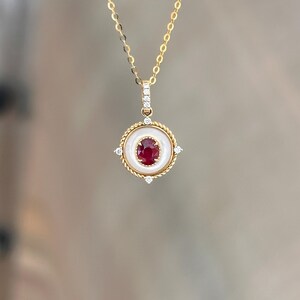 May include: A gold necklace with a pendant featuring a central red gemstone surrounded by a white circular border. The pendant is accented with small diamonds and a twisted gold rope design. The chain is delicate and gold.
