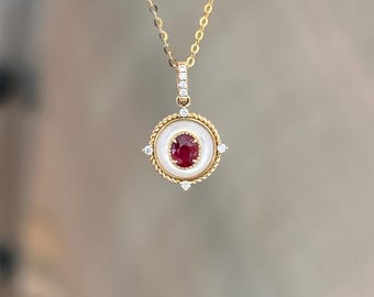Puede incluir: Un collar dorado con un colgante que presenta una gema roja central rodeada por un borde circular blanco. El colgante está adornado con pequeños diamantes y un diseño de cuerda dorada trenzada. La cadena es delicada y dorada.