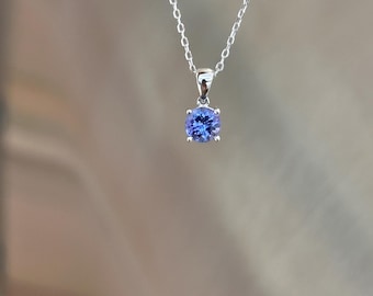 Puede incluir: Un collar de plata con un colgante de piedra preciosa azul redonda. La piedra preciosa está engastada en un engaste de cuatro puntas y cuelga de una delicada cadena. El colgante es de un color azul vibrante.