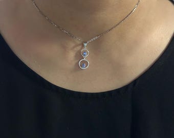 Puede incluir: Collar de plata con un colgante que presenta dos círculos abiertos, cada uno con una pequeña gema azul redonda. El collar se lleva sobre una parte superior negra, resaltando el diseño del colgante.
