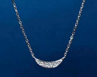 Puede incluir: Un collar plateado con un colgante de barra curvada engastado con pequeñas piedras preciosas transparentes. El colgante cuelga de una delicada cadena. El fondo es azul oscuro liso.