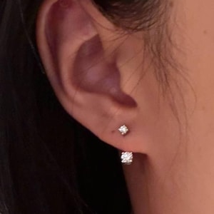 May include: A close-up of an ear with two small, sparkling diamond stud earrings. One earring is a single, small diamond, and the other is a larger, square-shaped cluster of diamonds. The earrings are set in a silver or white metal.
