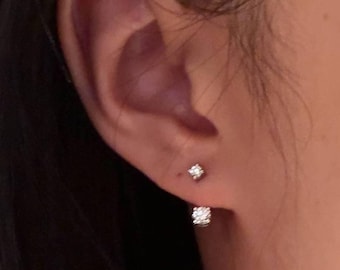 Puede incluir: Primer plano de una oreja con dos pendientes de diamantes pequeños y brillantes. Uno es un solo diamante pequeño, el otro es un grupo cuadrado más grande. Los pendientes están engastados en metal plateado o blanco.