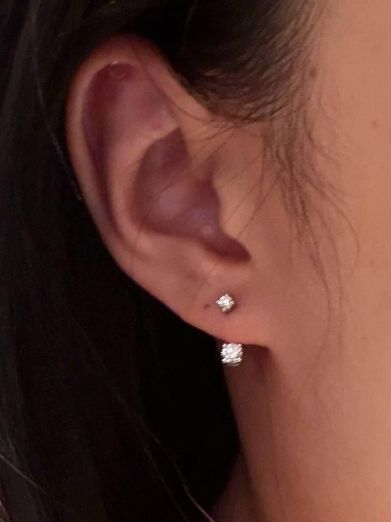 May include: A close-up of an ear with two small, sparkling diamond stud earrings. One earring is a single, small diamond, and the other is a larger, square-shaped cluster of diamonds. The earrings are set in a silver or white metal.