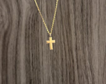 Puede incluir: Un delicado collar dorado con un pequeño colgante de cruz. La cruz tiene un diseño texturizado y cuelga de una fina cadena. El collar se muestra sobre un fondo oscuro y texturizado.