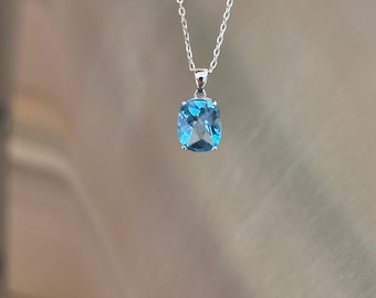 Puede incluir: Un collar de plata con un colgante cuadrado de piedra preciosa azul claro. El colgante es de un azul claro y vibrante, y la cadena es delicada. El collar se muestra sobre un fondo borroso y neutro.