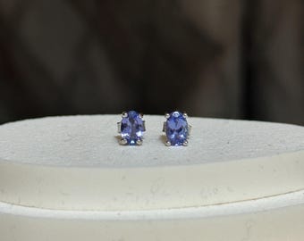 Puede incluir: Un par de pendientes de botón de tanzanita púrpura claro de forma ovalada. Las piedras preciosas están engastadas en plata u oro blanco. Los pendientes se exhiben sobre una superficie blanca con un fondo borroso.