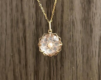 Puede incluir: Collar dorado con colgante redondo. El colgante tiene un centro claro y facetado con diseño de estrella y una pequeña perla en el centro. El colgante está rodeado por un marco dorado y festoneado. El collar cuelga de una cadena dorada.