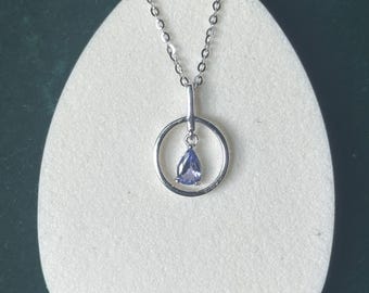 Puede incluir: Un collar de plata con un colgante de piedra preciosa en forma de lágrima, de color púrpura claro. El colgante está suspendido dentro de un círculo de plata, conectado a una fina cadena. El collar se exhibe sobre una superficie blanca ovalada.