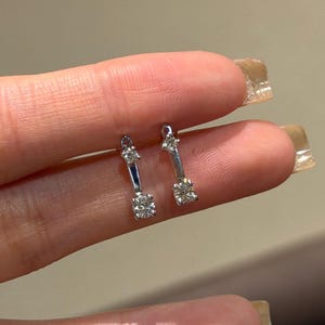 May include: A pair of silver-toned earrings. Each earring features a small, square-cut diamond at the bottom and a smaller diamond at the top, connected by a slender bar. The earrings are held up by a person's fingers.