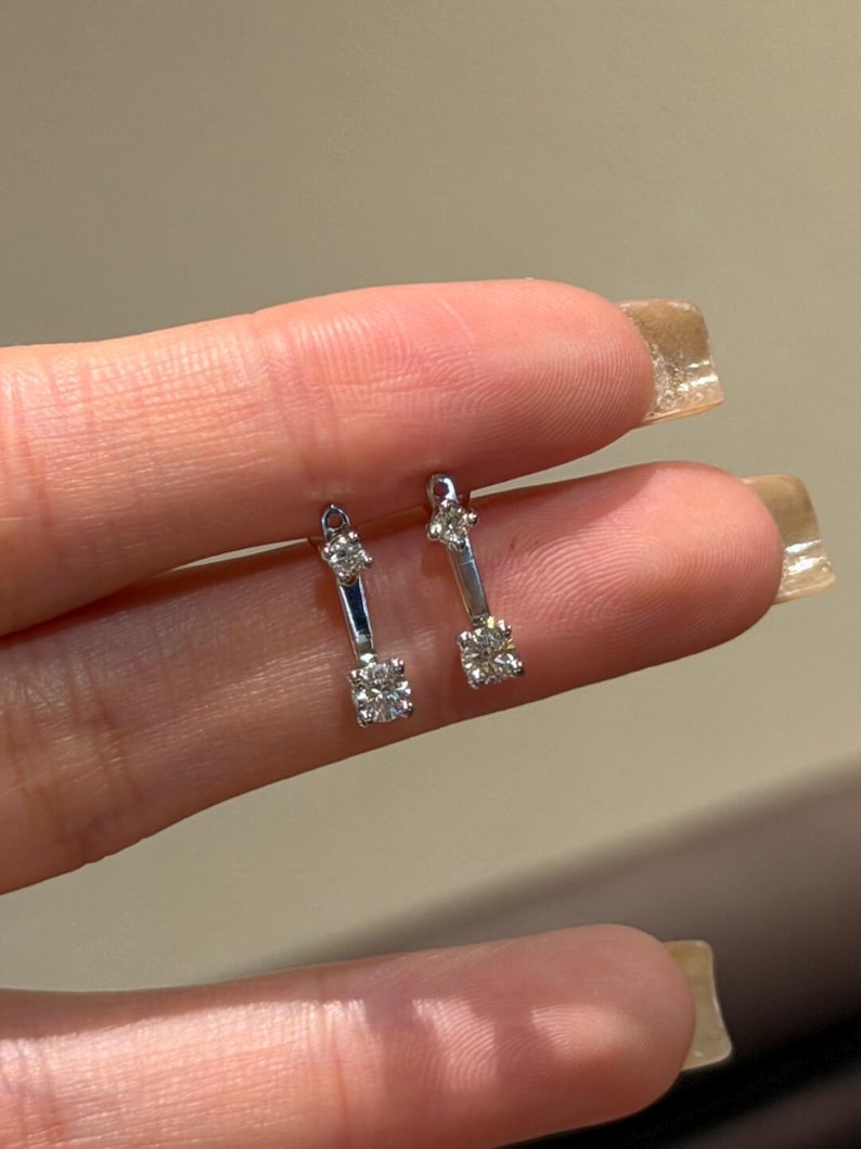 May include: A pair of silver-toned earrings. Each earring features a small, square-cut diamond at the bottom and a smaller diamond at the top, connected by a slender bar. The earrings are held up by a person's fingers.