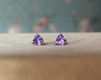Puede incluir: Un par de pendientes de botón con piedras preciosas moradas facetadas en forma de corazón. Los pendientes están engarzados en metal plateado y se muestran sobre una superficie neutra.