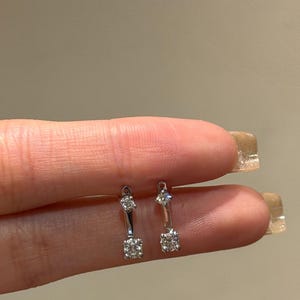 May include: A pair of silver earrings with a linear design. Each earring features a small, square cluster of clear stones at the bottom and a single, smaller stone at the top. The earrings are held between fingers against a neutral background.