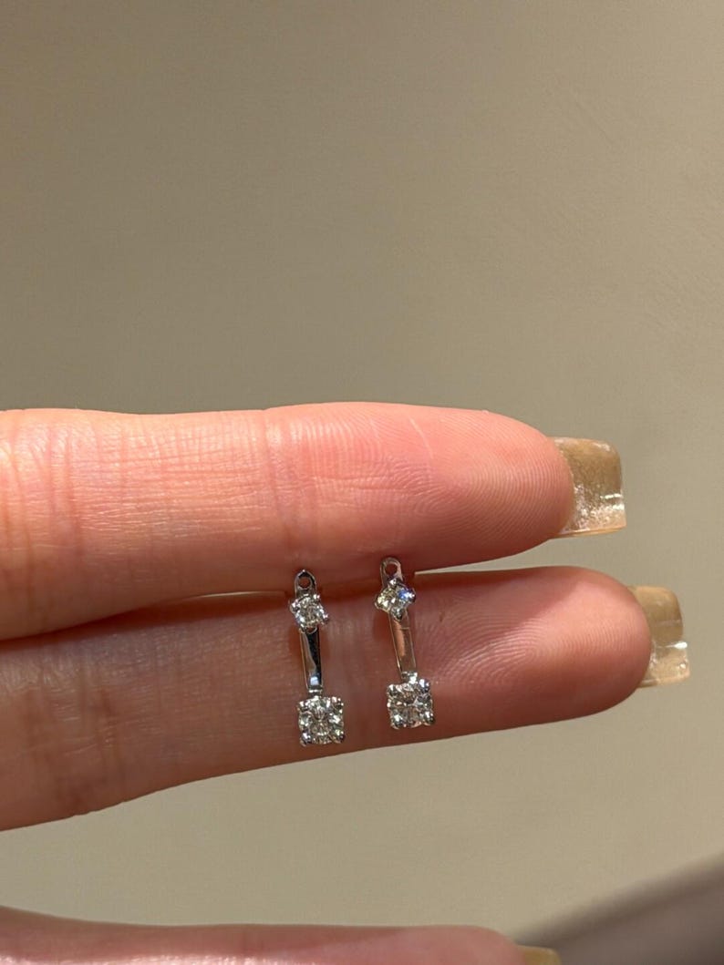 May include: A pair of silver earrings with a linear design. Each earring features a small, square cluster of clear stones at the bottom and a single, smaller stone at the top. The earrings are held between fingers against a neutral background.