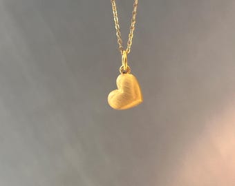Puede incluir: Un collar dorado con un colgante en forma de corazón. El colgante tiene una superficie texturizada y cuelga de una cadena delicada. El fondo es un degradado de gris y rosa.