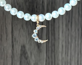 Puede incluir: Un delicado collar con una cadena de cuentas redondas e iridiscentes. Un colgante de media luna dorado, adornado con piedras preciosas azul claro y transparentes, cuelga del collar, creando un diseño celestial.