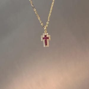 以下が含まれることがあります： ゴールドカラーのネックレス、クロス型ペンダント付き。クロスは赤い中心石と透明で輝く石で囲まれています。繊細なチェーンがニュートラルな背景に垂れ下がっています。