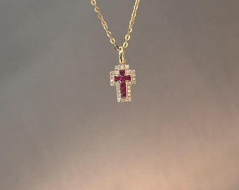 Puede incluir: Un collar dorado con un colgante de cruz. La cruz presenta una piedra central roja rodeada de piedras claras y brillantes. La delicada cadena cuelga sobre un fondo neutro.