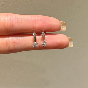 May include: A pair of silver earrings with a bar design. Each earring features a small, sparkling gemstone at the top and a larger, round gemstone at the bottom. The earrings are held up against a neutral background.