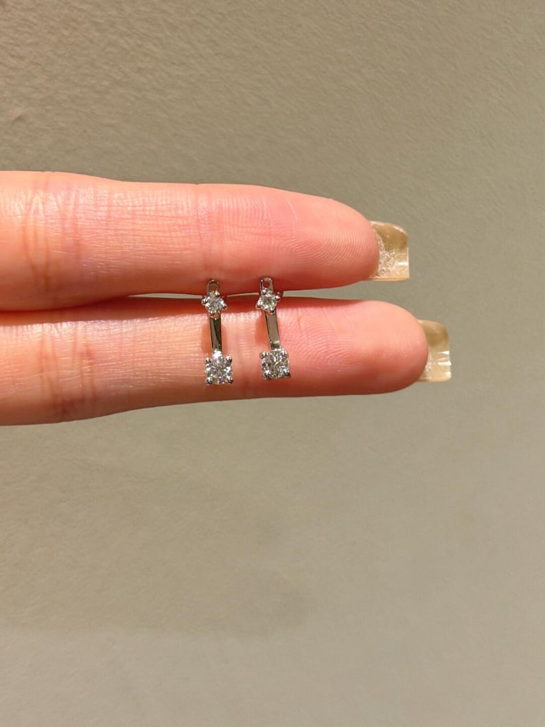 May include: A pair of silver earrings with a bar design. Each earring features a small, sparkling gemstone at the top and a larger, round gemstone at the bottom. The earrings are held up against a neutral background.