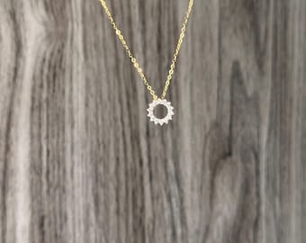 Puede incluir: Delicado collar dorado con un colgante con un círculo de pequeñas piedras blancas brillantes. El collar tiene una fina cadena dorada y se muestra sobre un fondo de madera.