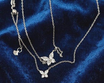Puede incluir: Collares colgantes de mariposa plateados con piedras claras y brillantes. Los collares son superpuestos, uno de ellos con un pequeño dije en forma de corazón. Las joyas se exhiben sobre una superficie de terciopelo azul oscuro.