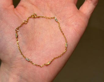 Puede incluir: Una delicada pulsera dorada con pequeñas gemas brillantes espaciadas uniformemente a lo largo de la cadena. La pulsera se muestra en la palma de una mano abierta, mostrando sus finos detalles y su elegante diseño. El cierre es visible.