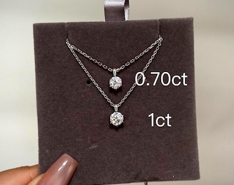 Puede incluir: Dos collares plateados con piedras preciosas redondas y transparentes se exhiben sobre un fondo de terciopelo marrón oscuro. El collar superior tiene una gema de 0,70 ct, y el collar inferior tiene una gema de 1 ct.