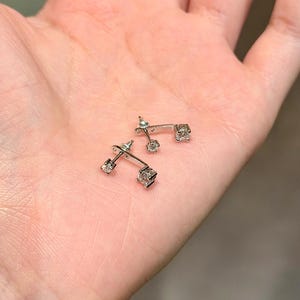 May include: A pair of silver-toned earrings with a square, clear gemstone at each end. The earrings are small and delicate, with a minimalist design. The earrings are displayed on a light-colored surface.