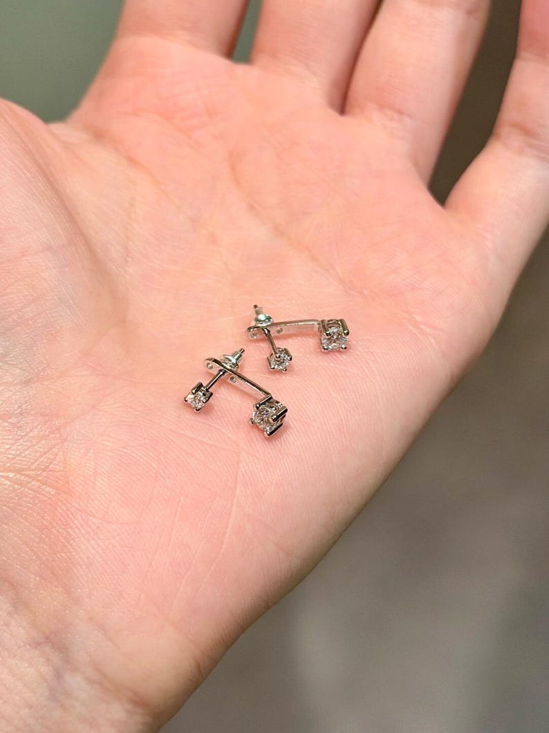 May include: A pair of silver-toned earrings with a square, clear gemstone at each end. The earrings are small and delicate, with a minimalist design. The earrings are displayed on a light-colored surface.