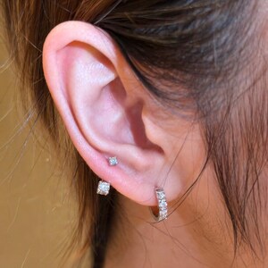 May include: Close-up of an ear adorned with three different earrings. The ear has a small, round, light blue stud, a square-shaped stud, and a small hoop earring with multiple clear stones.