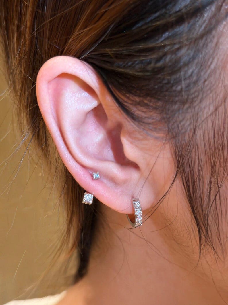 May include: Close-up of an ear adorned with three different earrings. The ear has a small, round, light blue stud, a square-shaped stud, and a small hoop earring with multiple clear stones.
