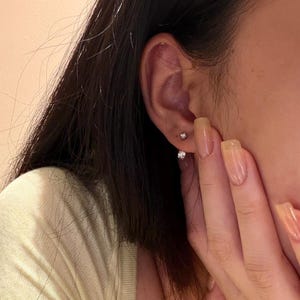 May include: Close-up of a person's ear adorned with two small, sparkling stud earrings. The earrings appear to be made of a shiny metal, possibly silver or white gold, and feature a single, clear gemstone. The person's hand is visible.