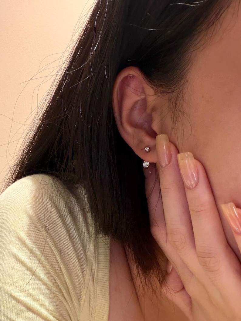 May include: Close-up of a person's ear adorned with two small, sparkling stud earrings. The earrings appear to be made of a shiny metal, possibly silver or white gold, and feature a single, clear gemstone. The person's hand is visible.