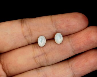 Puede incluir: Un par de pendientes de botón ovalados de piedra preciosa blanca iridiscente con engastes dorados. Los pendientes se muestran sobre un fondo oscuro.