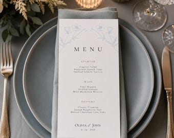 May include: A formal table setting with a menu card. The menu card is white with a floral design and the word "MENU" at the top. The menu lists starter, main, and dessert courses. The plates are gray, and the silverware is gold.