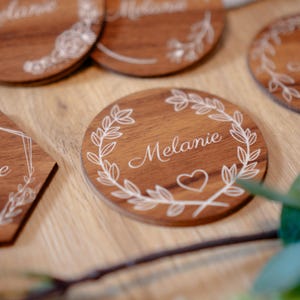 Può includere: Sottobicchieri in legno con il nome "Melanie" inciso in corsivo. I sottobicchieri sono rotondi ed esagonali, con un disegno floreale bianco attorno al nome. Un cuore è inciso su un sottobicchiere.