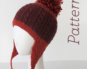 Puede incluir: Un gorro de punto marrón y naranja con un pompón en la parte superior y orejeras que se atan debajo de la barbilla. El gorro está hecho con un patrón de punto de cable.