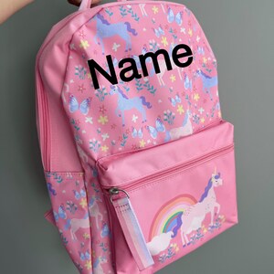 Peut inclure: Sac à dos rose avec un motif de licorne et d'arc-en-ciel sur la poche avant. Le sac à dos présente un motif de licornes, de papillons et de fleurs. Le mot "Name" est imprimé sur le devant.