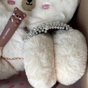 Puede incluir: Un art&iacute;culo blanco con forma de oso de peluche con ojos cerrados, detalles brillantes y un coraz&oacute;n dorado. Se ve un bol&iacute;grafo marr&oacute;n con una figura de oso y las palabras "WONDERFUL LIFE". Tambi&eacute;n hay una diadema adornada con perlas.