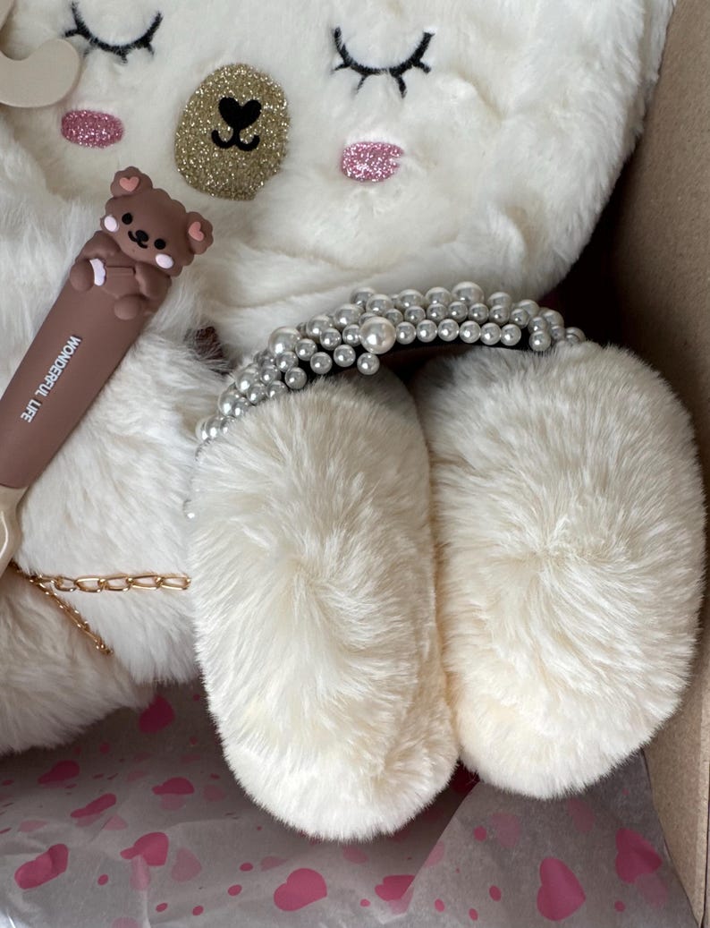 Puede incluir: Un art&iacute;culo blanco con forma de oso de peluche con ojos cerrados, detalles brillantes y un coraz&oacute;n dorado. Se ve un bol&iacute;grafo marr&oacute;n con una figura de oso y las palabras "WONDERFUL LIFE". Tambi&eacute;n hay una diadema adornada con perlas.