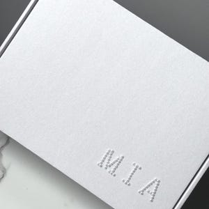 Puede incluir: Una caja de regalo blanca texturizada con el nombre "MIA" en relieve en la parte delantera. La caja est&aacute; cerrada y tiene un dise&ntilde;o limpio y minimalista. La caja est&aacute; sobre una superficie blanca y gris.