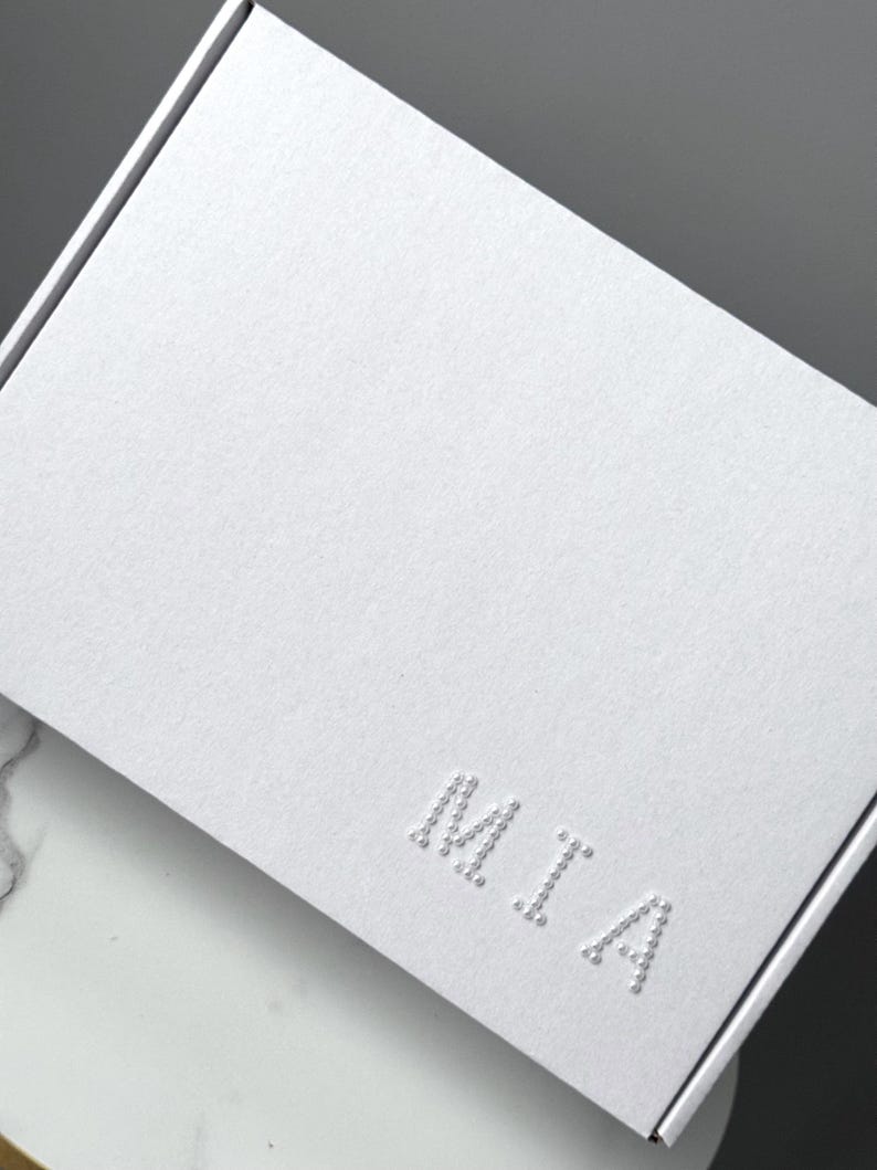 Puede incluir: Una caja de regalo blanca texturizada con el nombre "MIA" en relieve en la parte delantera. La caja est&aacute; cerrada y tiene un dise&ntilde;o limpio y minimalista. La caja est&aacute; sobre una superficie blanca y gris.