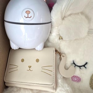 Puede incluir: Un humidificador blanco con forma de oso con detalles en oro rosa, una cartera crema con cara de gato y una mochila de unicornio blanca y esponjosa se exhiben dentro de una caja. La cartera tiene bigotes y ojos dorados. El unicornio tiene una nariz de purpurina rosa.