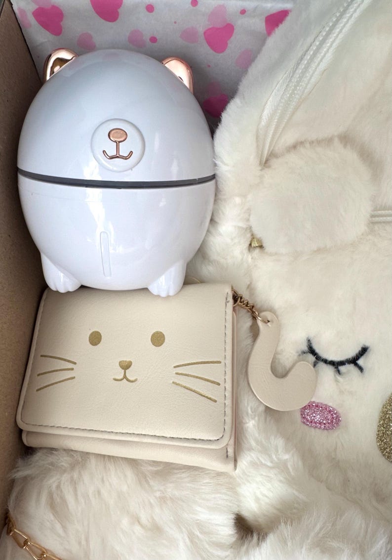 Puede incluir: Un humidificador blanco con forma de oso con detalles en oro rosa, una cartera crema con cara de gato y una mochila de unicornio blanca y esponjosa se exhiben dentro de una caja. La cartera tiene bigotes y ojos dorados. El unicornio tiene una nariz de purpurina rosa.