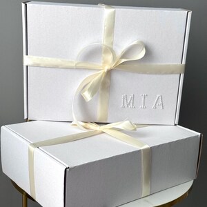 Puede incluir: Dos cajas de regalo blancas apiladas, atadas con cintas y lazos color crema. La caja superior tiene las letras "MIA" en relieve. Est&aacute;n sobre una peque&ntilde;a mesa dorada.