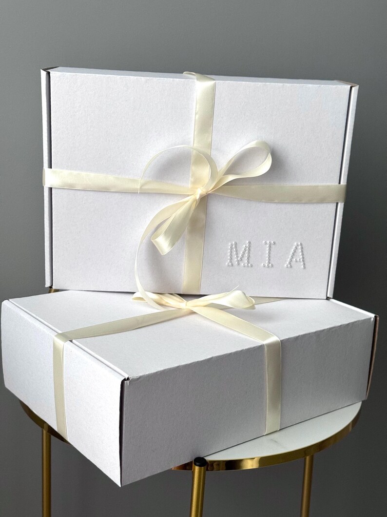 Puede incluir: Dos cajas de regalo blancas apiladas, atadas con cintas y lazos color crema. La caja superior tiene las letras "MIA" en relieve. Est&aacute;n sobre una peque&ntilde;a mesa dorada.