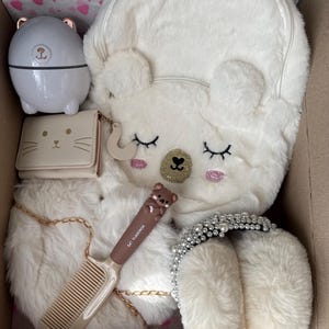 Puede incluir: Una colecci&oacute;n de accesorios de felpa blanca, que incluye una mochila con forma de oso, una peque&ntilde;a cartera con cara de gato, un peine y una diadema con perlas. Tambi&eacute;n hay un peque&ntilde;o recipiente blanco con tem&aacute;tica de oso.