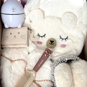 Puede incluir: Una colecci&oacute;n de accesorios blancos y crema, que incluyen una mochila con forma de oso, una cartera con cara de gato, un peine con un adorno de oso, una diadema de perlas y un peque&ntilde;o humidificador blanco. Los art&iacute;culos est&aacute;n dispuestos en una caja.